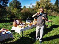 A man is standing in a meadow eating an apple. Behind him are tables with cloths and baskets full of apples and bread. It is a sunny day and there are trees in the background.