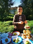 A person stands in front of an outdoor table on which plates with sliced apples and glasses with apple slices are presented. A sunny garden can be seen in the background.