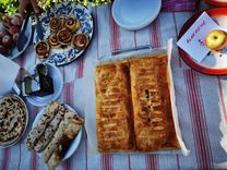 A table with a red and white striped tablecloth and various baked goods: A large apple strudel, plates of cinnamon buns, apple pie and a glass of dark liquid.