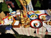 An outdoor table with various apple desserts, surrounded by people. In the center is a carafe holder with apples, flanked by plates with apple pie and apple roses.