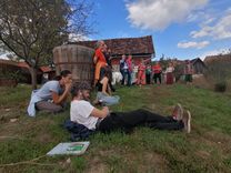 The group stands in front of a house with a tiled roof, listening to a lecture as part of a seed workshop. Two people are sitting comfortably on the ground in the foreground. It is a sunny day with some clouds in the sky.