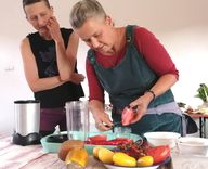 Two women are in the kitchen; one is chopping vegetables while the other watches. Various types of vegetables are spread out on the table, and a blender is ready for use.