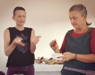 Two people are standing together, one holding something in a bowl while the other gestures with their hands.