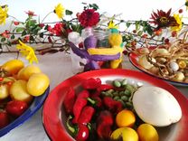 Colorful plates with various fruits and vegetables are lying on a table. One plate holds bell peppers and small tomatoes. Another plate has yellow fruits, likely lemons. In the background, a vase is decorated with colorful flowers and branches, creating an autumnal atmosphere.